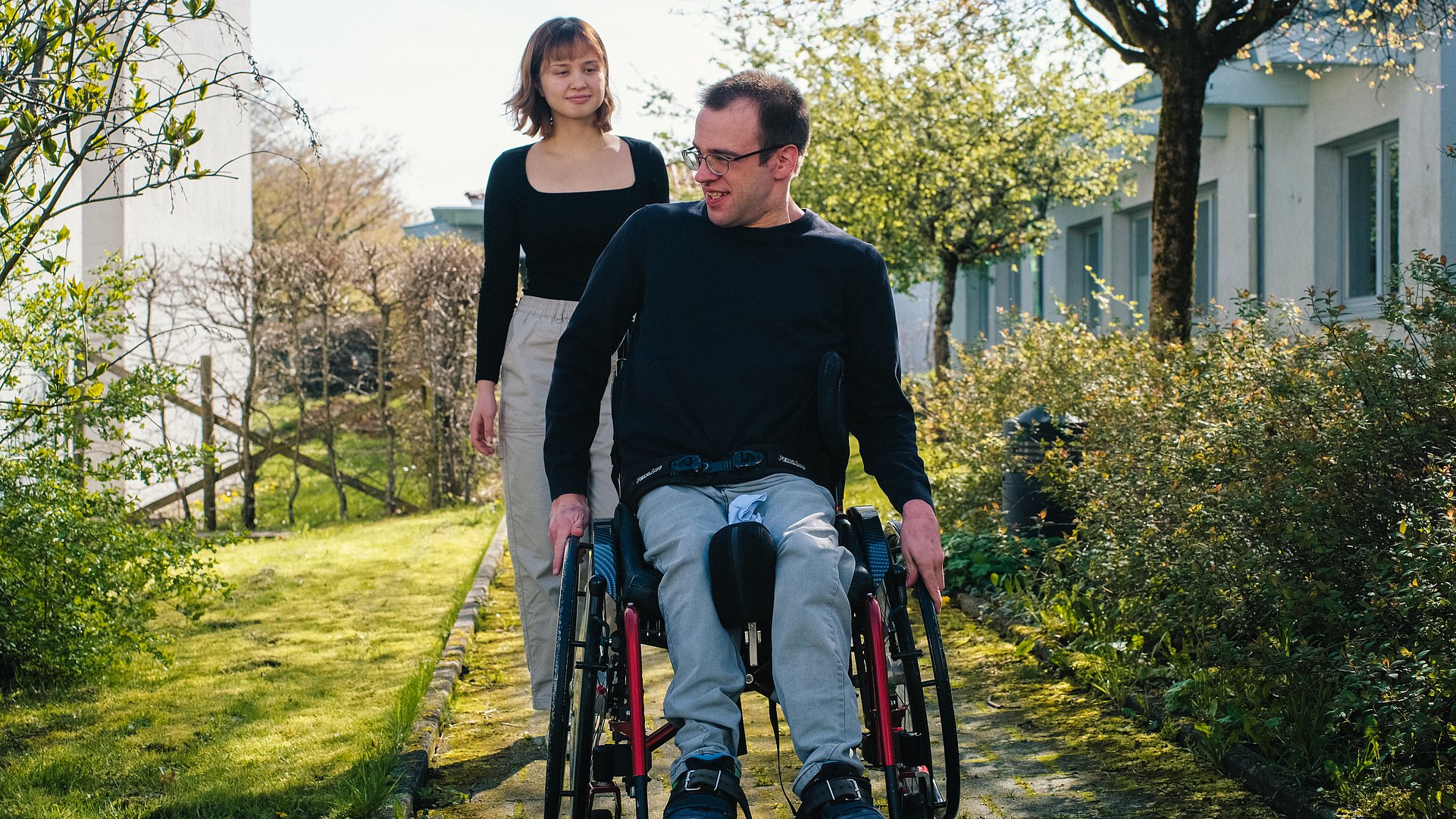 Ein Mann, der einen Rollstuhl nutzt und eine junge Frau sind draußen im Freien auf einem Weg. 