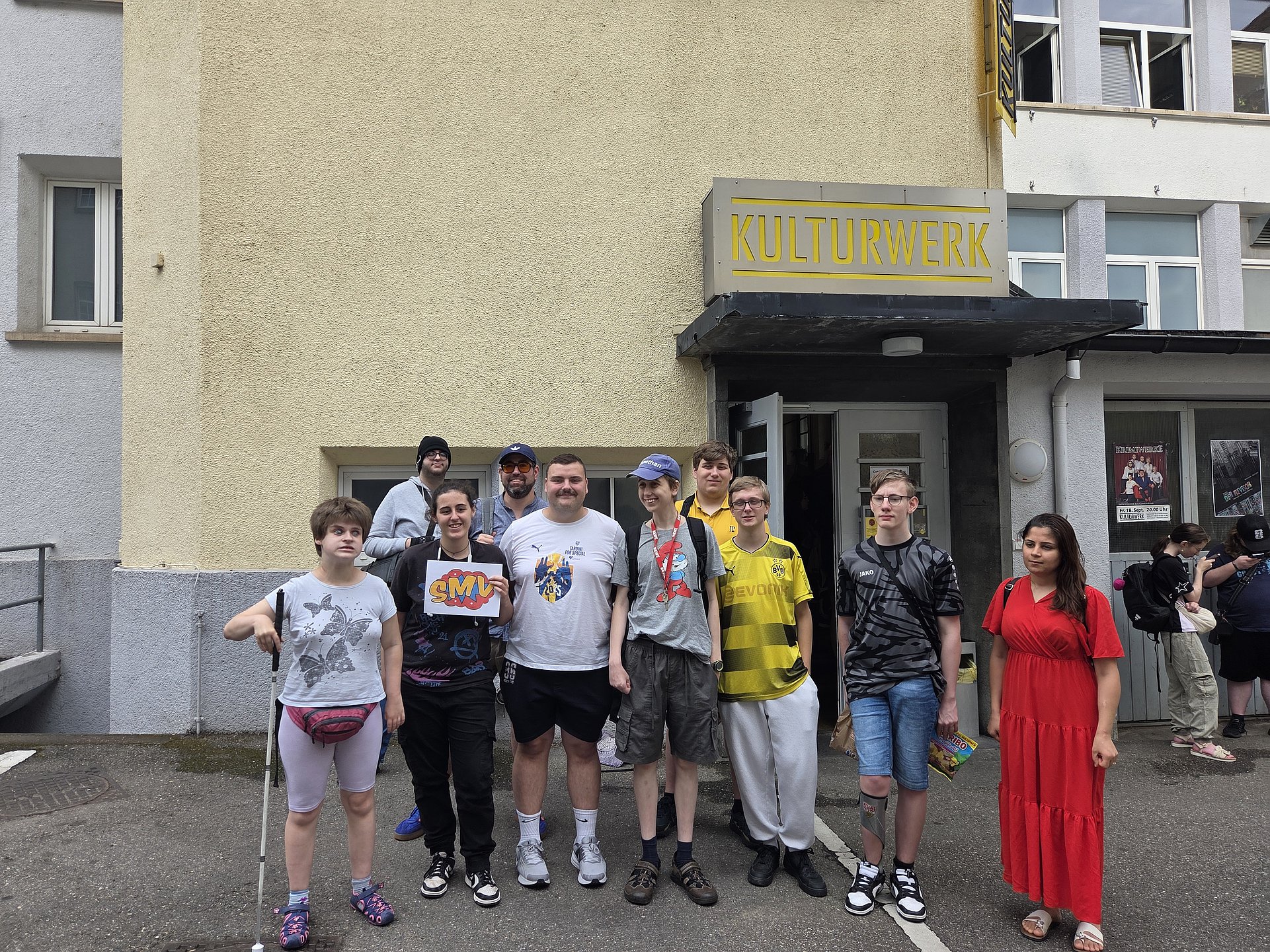 Eine Gruppe von zwölf Personen steht vor einem Gebäude mit dem Schild „KULTURWERK“.  