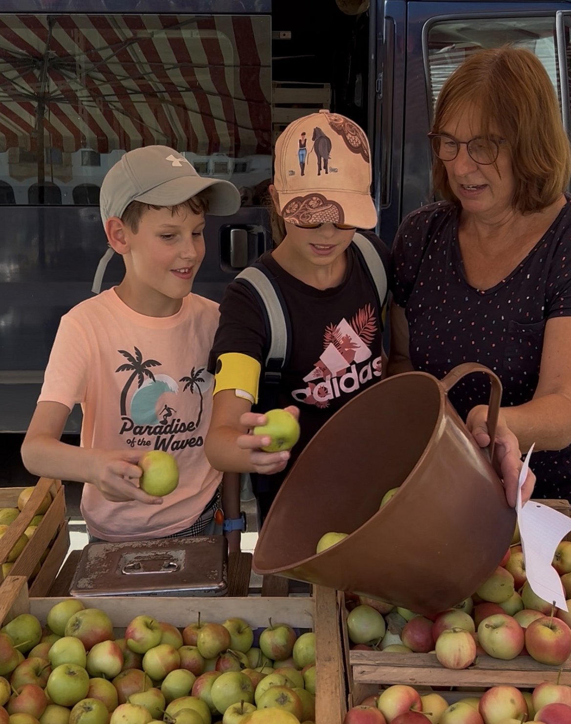 Zwei Kinder und eine Frau stehen an einem Marktstand und füllen Äpfel in ein braunes Gefäß aus Kunststoff