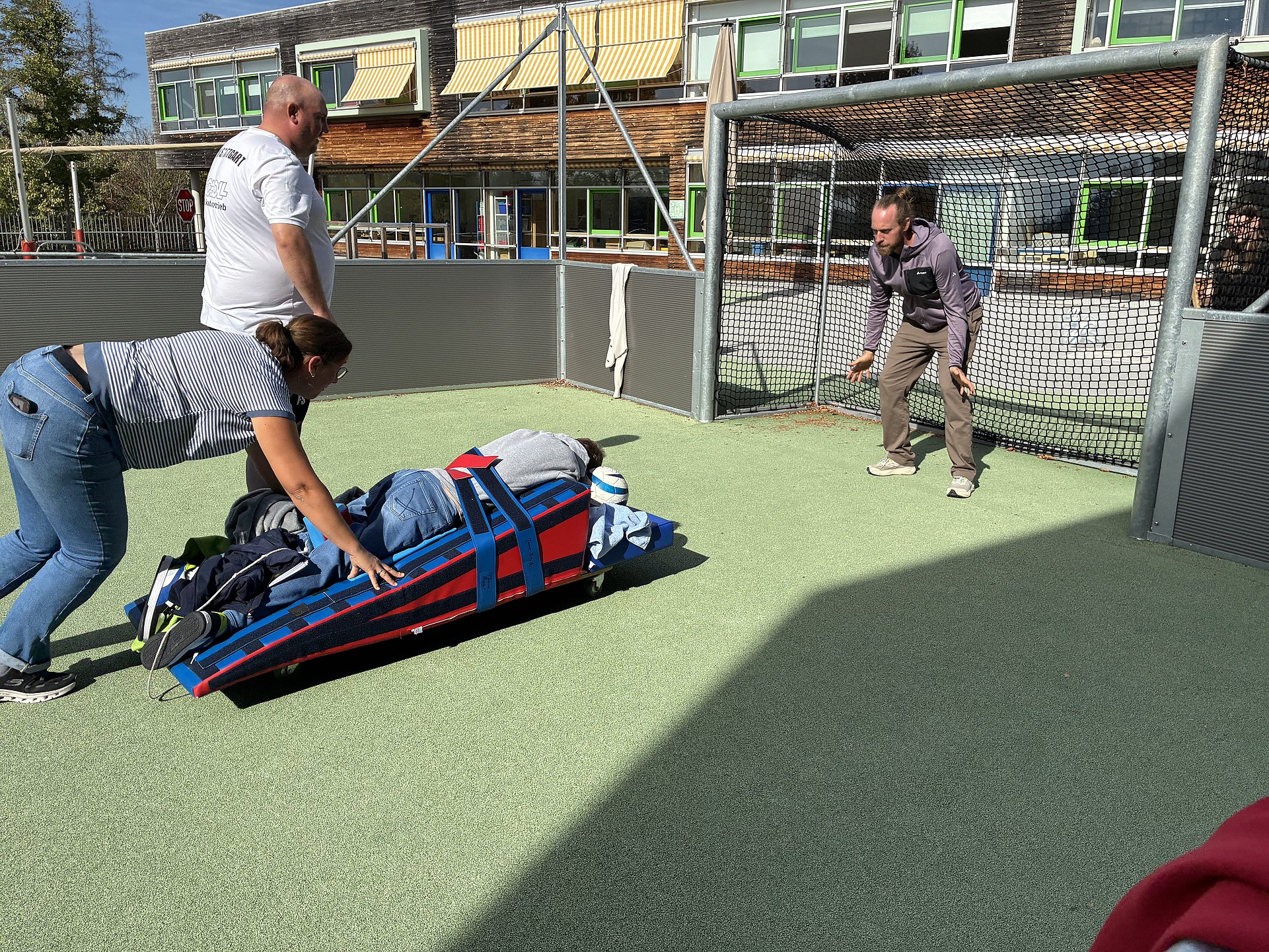 Ein kleiner Sportplatz mit einem Tor im Freien. Links im Vordergrund liegt eine rote und blaue Rolltrage oder ein flacher Wagen auf dem Boden, darauf liegt eine Person vollständig ausgestreckt auf dem Bauch, mit Jeans und Jacke bekleidet, die Füße ragen leicht über das Ende hinaus. Eine andere erwachsene Person in enger Jeans und gestreiftem T‑Shirt beugt sich weit nach vorn und schiebt die Trage kräftig nach rechts in Richtung Tor. Hinter ihr steht ein weiterer Erwachsener mit Glatze und Bart, weißem T‑Shirt und dunkler Hose, mit leicht vorgeneigtem Oberkörper, als würde er die Situation beobachten oder begleiten. Rechts vor dem Tor steht ein Mann mit mittlerer Hautfarbe, dunklem, nach hinten gebundenem Haar, grauer Kapuzenjacke und beigefarbener Hose in einer leicht geduckten, breiten Abwehrhaltung, die Arme seitlich angewinkelt, als wolle er das „Gefährt“ wie einen Ball abwehren.