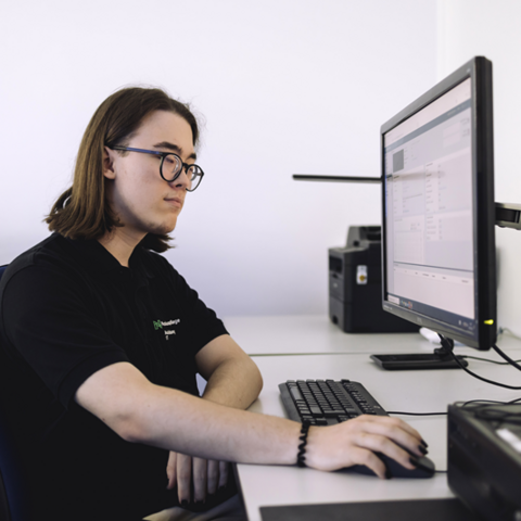 Ein junger Mann in einem schwarzen T-Shirt sitzt an einem Schreibtisch. Er hat schulterlange braune Haare und trägt eine Brille. Mit seiner rechten Hand bedient er eine Maus. Er blickt auf einen Computerbildschirm