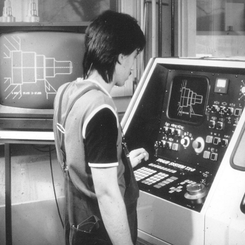Eine schwarz weiß Fotografie. Ein Mann steht vor einem elektronischen Gerät in der Werkstatt und blickt auf den Bildschirm
