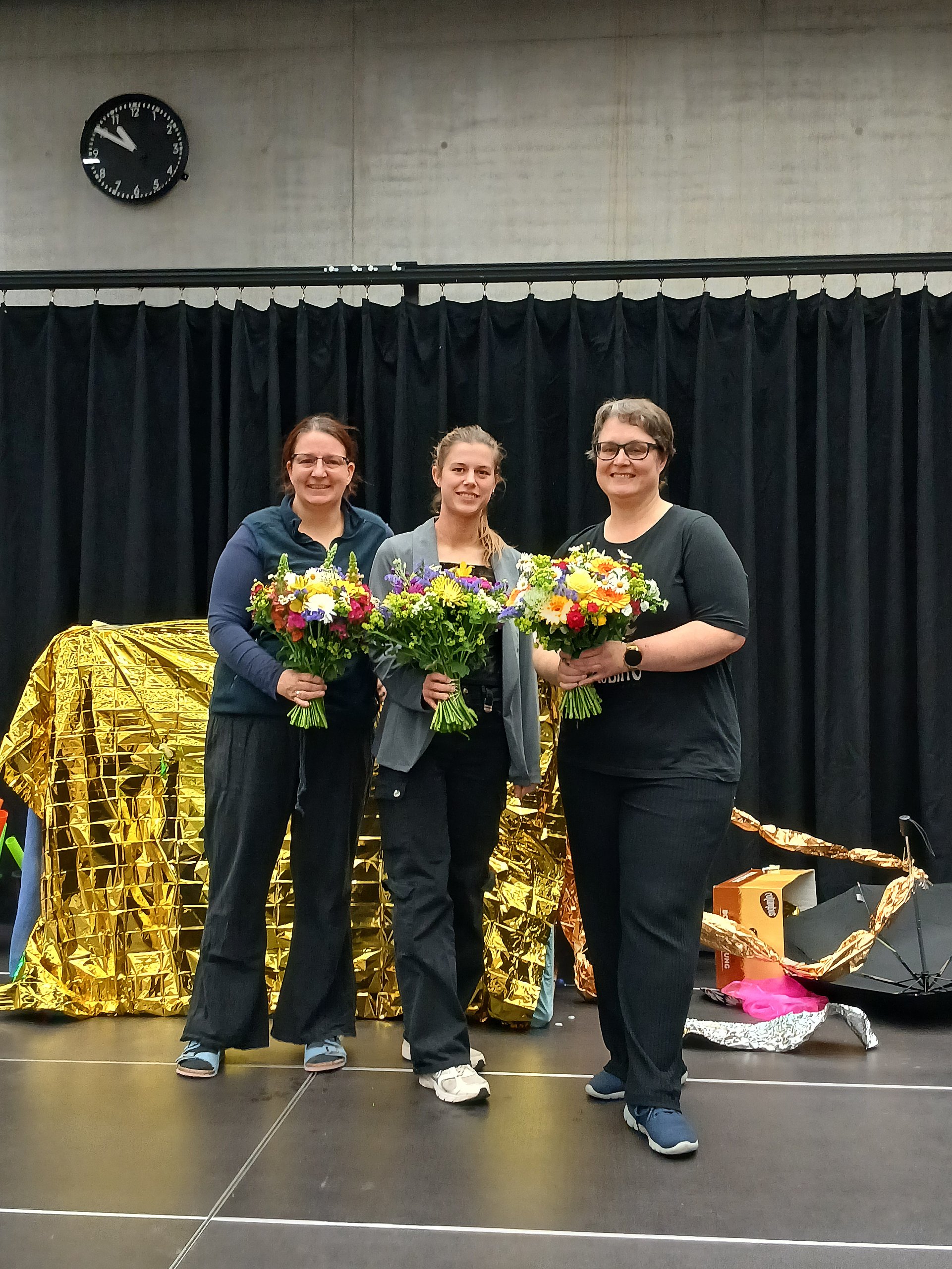 Drei Frauen stehen nebeneinander auf einer Bühne und halten bunte Blumensträuße in den Händen.   Alle drei lächeln in Richtung Kamera.   Die Frau links trägt eine dunkle Hose, ein dunkelblaues Oberteil mit Kapuze und helle Sportschuhe; sie hat hellen Hautton und dunkle, zurückgebundene Haare.   Die Frau in der Mitte hat hellen Hautton, mittelblonde, nach hinten gebundene Haare, eine dunkle Hose, ein dunkles Oberteil und darüber eine offene, hellere Jacke sowie helle Sportschuhe.   Die Frau rechts ist etwas kräftiger gebaut, hat hellen Hautton, kurze dunkle Haare, eine dunkle Hose, ein dunkles T‑Shirt und dunkelblaue Schuhe; sie trägt eine Armbanduhr am linken Handgelenk.   Die Blumensträuße sind groß und gemischt, mit vielen Farben wie Gelb, Weiß, Lila, Rot und Grün.   Hinter den Frauen hängt ein schwarzer Vorhang, davor liegt links eine große, goldglänzende Decke oder Folie, die gefaltet und drapiert ist.