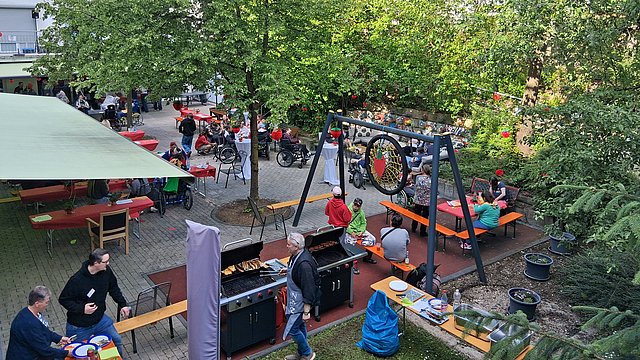 Der Außenbereich des Pau und Charlotte Kniese Hauses. Es sind Biertische und Bierbänke aufgestellt. Die Bäume haben grüne Blätter, der Himmel ist blau. Man sieht viele Menschen. 