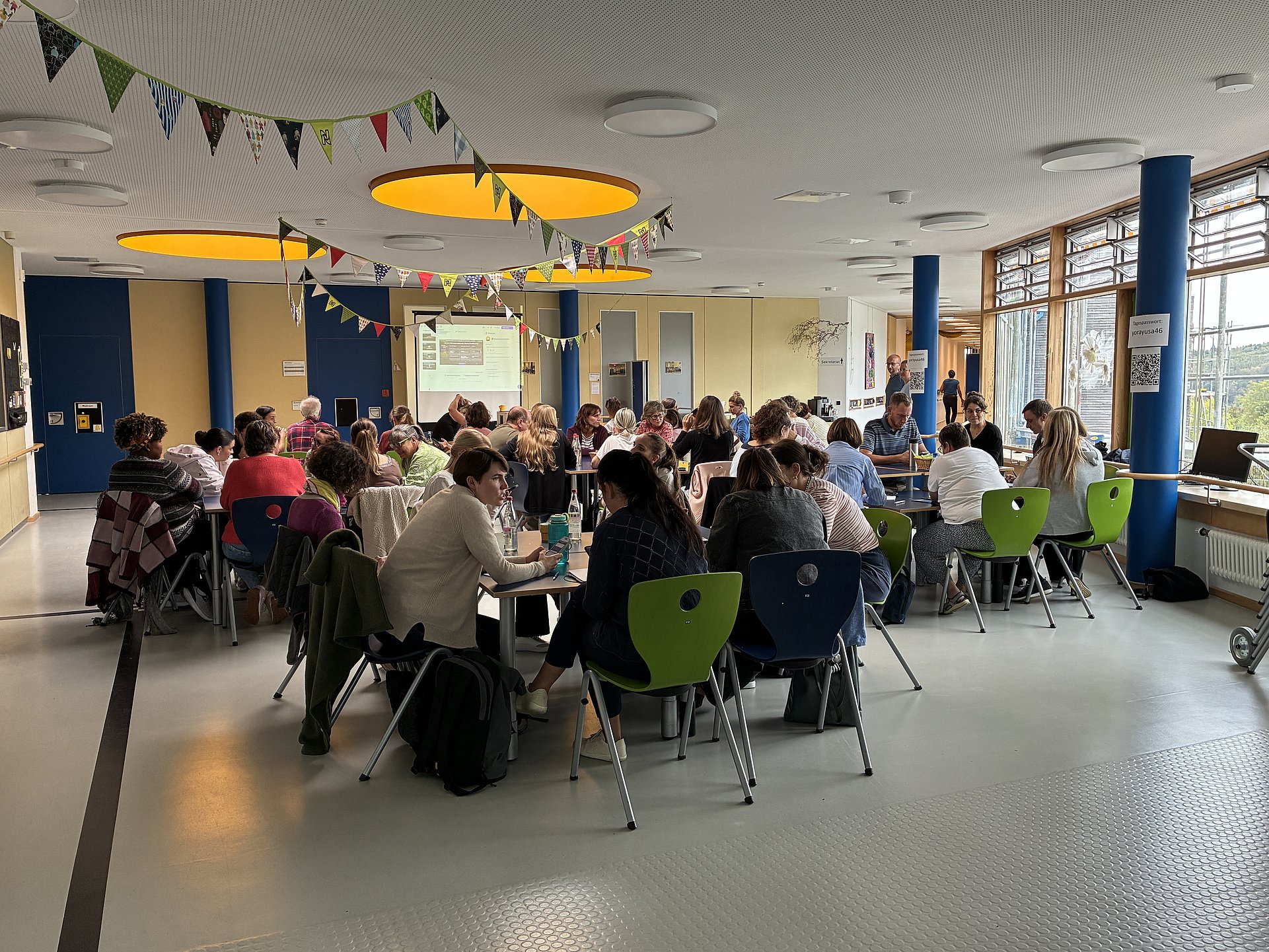 In einem hellen großen Raum sitzen viele Personen gemeinsam an zusammengestellten Tischen und unterhalten sich. An der Decke sind bunte Wimpel aufgehängt