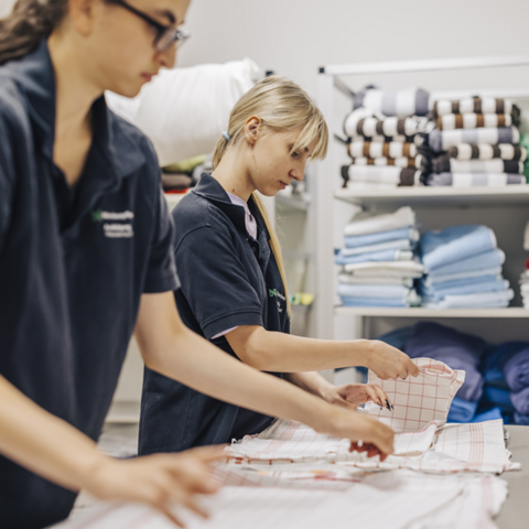 Zwei junge Frauen stehen nebeneinander vor einem Tisch. Darauf falten sie Küchentücher