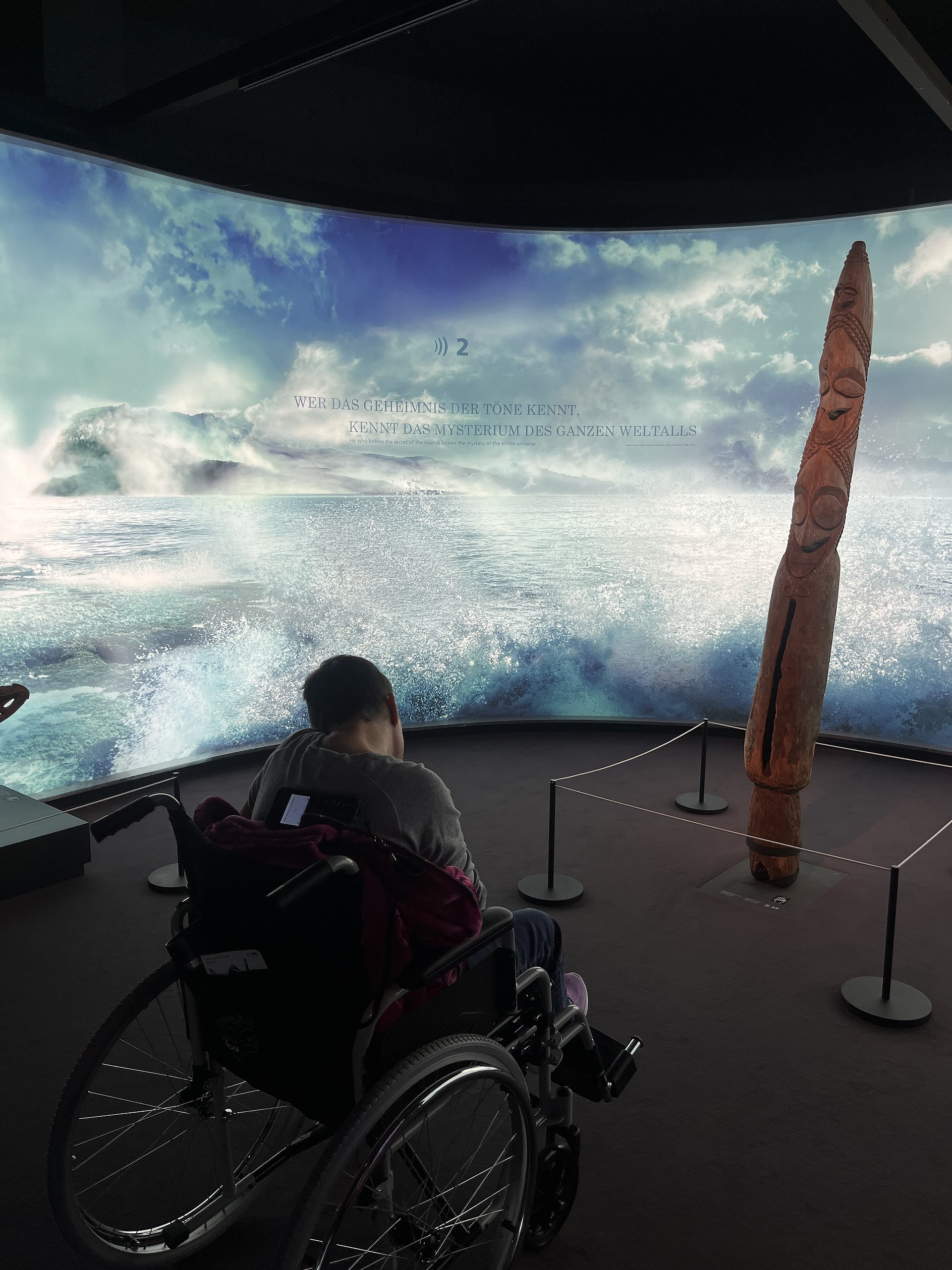 Eine Person im Rollstuhl in einem Ausstellungsraum vor einer großen, gebogenen Projektionswand mit Meeresszene. Die Projektionswand zeigt hohe, aufschäumende Wellen in Türkis- und Blautönen, darüber teils wolkiger Himmel. Im oberen mittleren Bereich der Wand steht Text; darüber ein Lautsprecher‑Symbol mit der Zahl 2. Der lesbare Teil des Textes lautet: „WER DAS GEHEIMNIS DER TÖNE KENNT, KENNT DAS MYSTERIUM DES GANZEN WELTALLS“. Rechts im Bild steht eine hohe, senkrechte Holzskulptur mit eingeschnitzten Mustern, die von vier Absperrpfosten mit Seilen begrenzt wird.