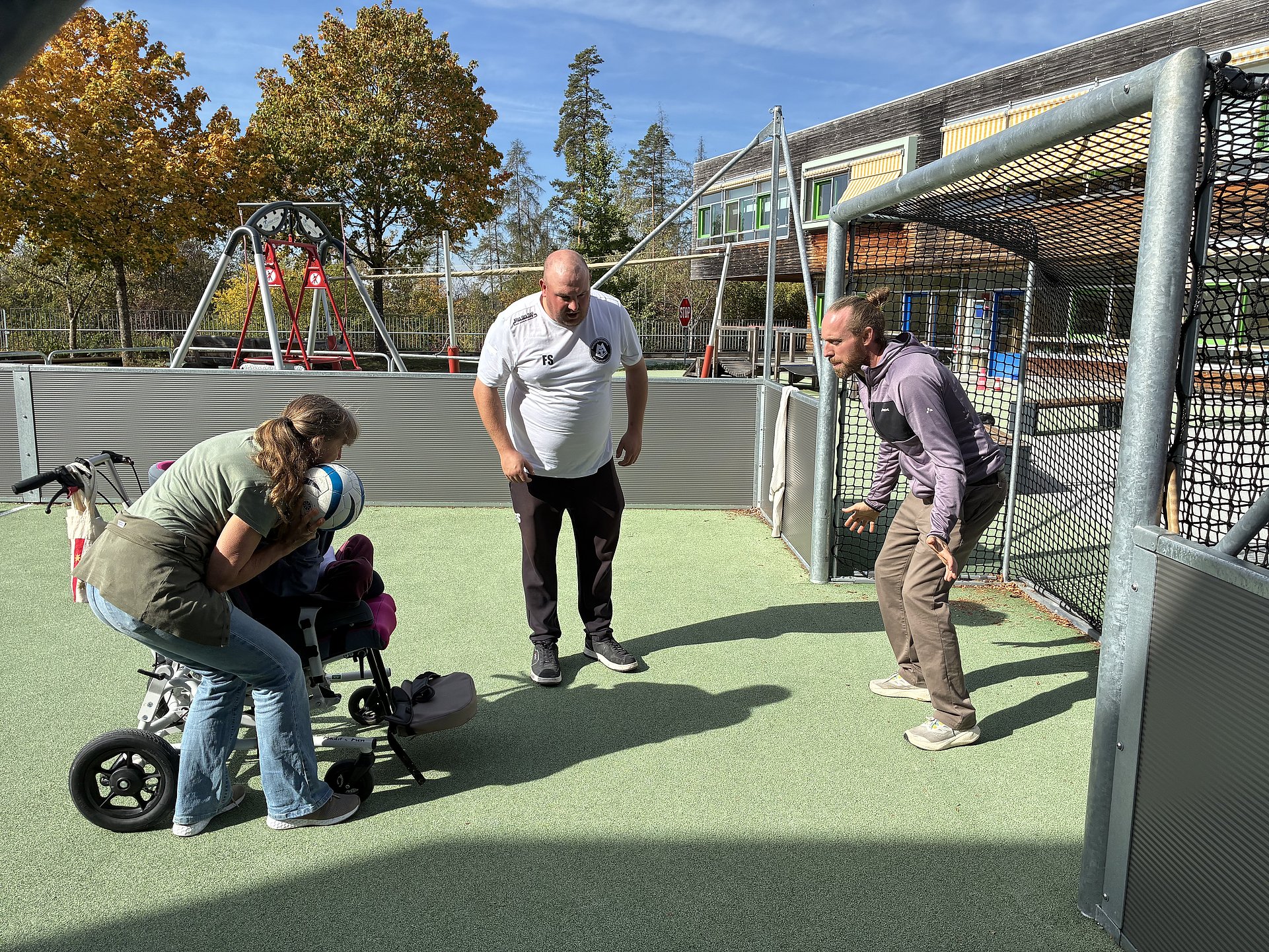 Drei Erwachsene und ein Kind auf einem kleinen Fußballfeld mit Tor. Links steht eine Frau mit hellbraunen, schulterlangen Haaren, die nach hinten zusammengebunden sind. Sie trägt ein olivgrünes T‑Shirt, eine beige Schürze oder Weste und eine hellblaue Jeans mit weißen Sportschuhen. Vor ihr ist ein spezieller Kinderrollstuhl mit drei Rädern und seitlicher Stützkonstruktion. In diesem sitzt ein Kind mit pinkfarbenen Polstern am Sitz und an den Fußstützen. Die Frau beugt den Oberkörper nach vorn über das Kind und hält einen weiß‑blauen Fußball direkt vor das Gesicht oder den Oberkörper des Kindes, als ob sie beim Schießen oder Passen helfen würde.