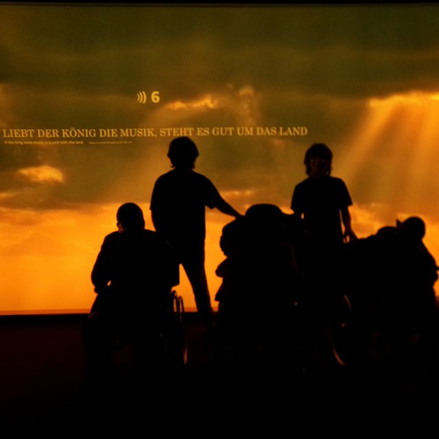 Schattenrisse mehrerer Menschen vor einem intensiv orange-gelben Sonnenuntergang.   Im Hintergrund sind dramatische Wolken und Sonnenstrahlen, die von rechts oben schräg nach unten fallen.   Vorne zeichnen sich deutlich dunkle Umrisse von mindestens vier Personen ab, einige davon wirken so, als säßen sie in Rollstühlen oder auf Rollbrettern, andere stehen oder hocken daneben.   Links oben steht in großer weiß-gelber Schrift: „LIEBT DER KÖNIG DIE MUSIK, STEHT ES GUT UM DAS LAND“.  