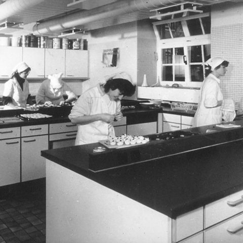 Eine schwarz weiß Fotografie. In einer Küche arbeiten junge Frauen an zwei Kochinseln. Sie tragen Arbeitskleidung mit einer Kopfbedeckung und bereiten das Essen zu