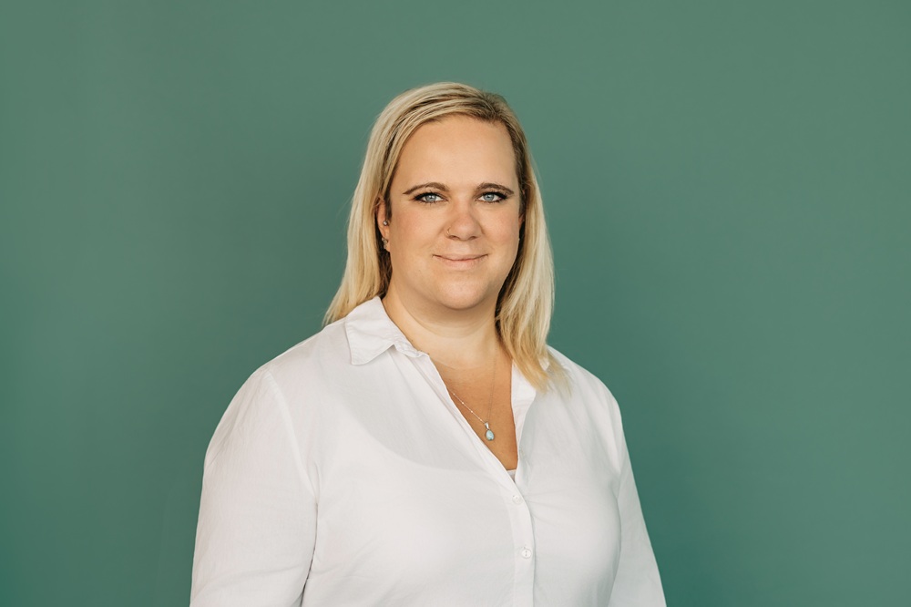 Eine Frau steht vor einem grünem Hintergrund. Sie trägt eine weiße Bluse. Sie hat schulterlange blonde glatte Haare.  