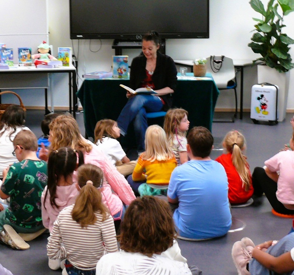 Eine Gruppe Kinder sitzt auf dem Boden des Klassenzimmers. Vor ihnen sitzt eine Frau auf einem Stuhl und liest aus einem Buch vor.