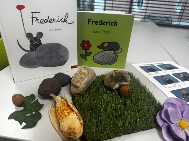 Auf einem Tisch stehen zwei Bilderbücher die eine Maus zeigen und den Titel Frederick tragen. Vor den Büchern liegen Steine, Blätter, ein Maiskolben und ein rechteckiges Stück Kunstrasen. Darauf sitzt eine kleine Plüschmaus, die eine Walnuss hält.