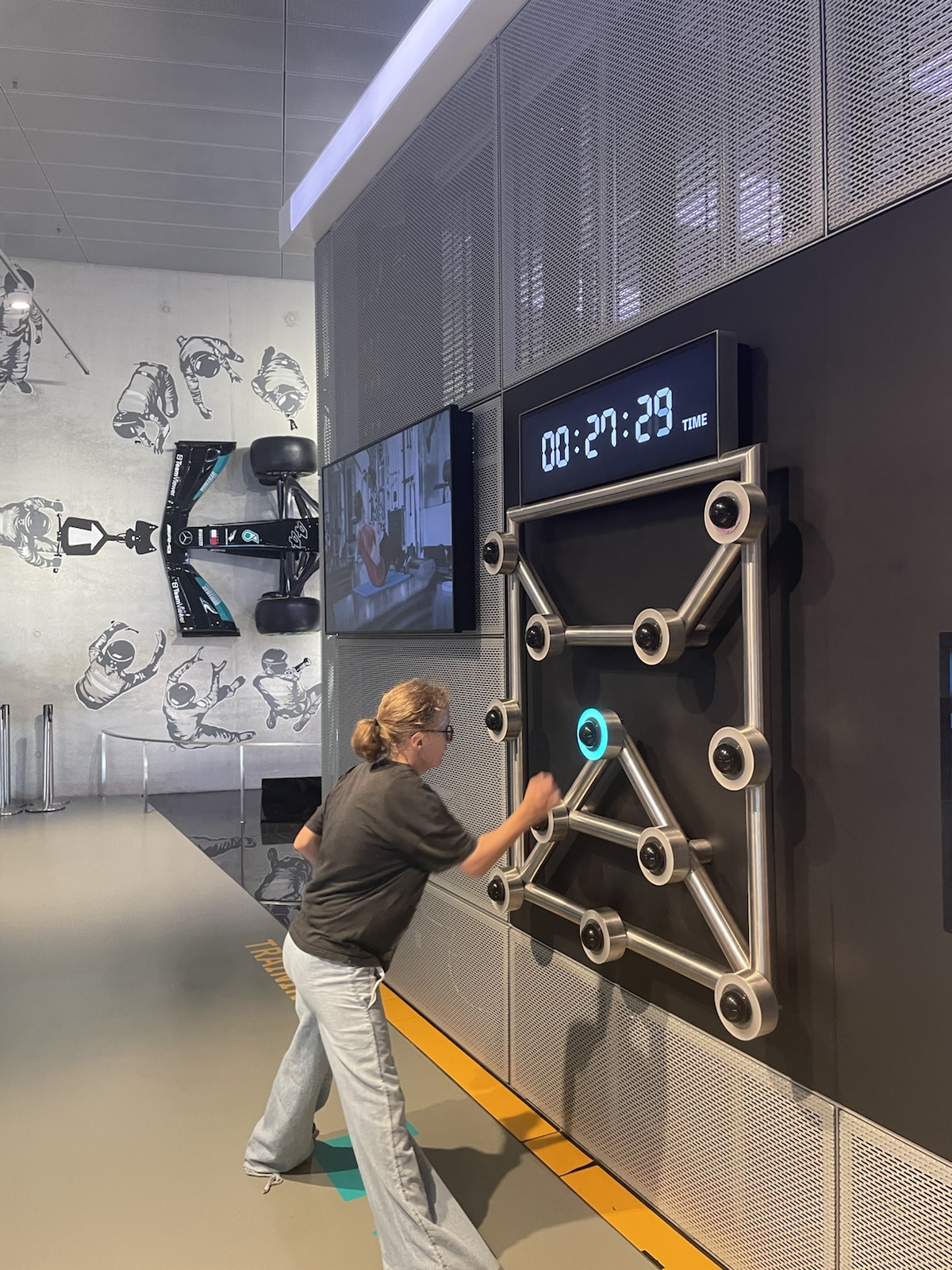 Eine Mädchen mit einem blonden Pferdeschwanz und einer Brille steht in einem Museum vor einer großen dunklen Wand. And der Wand sind runde metallische Objekte befestigt. Eines davon leuchtet blau auf. Die Objekte sind untereinander mit metallischen Leisten verbunden. Das Mädchen drückt mit ihrer rechten Hand einer der Objekte
