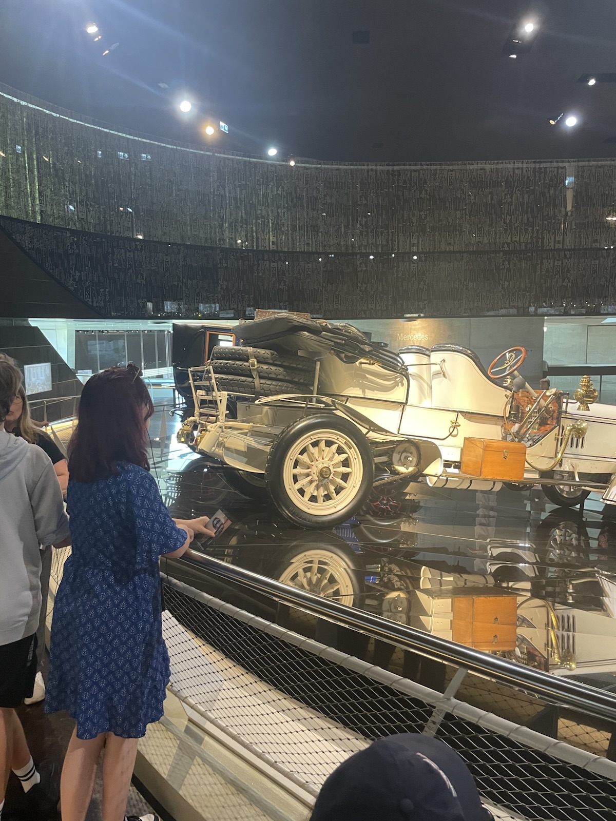 Eine Frau steht in einem Museumsraum. Sie trägt ein blaues Kleid und Schulterlange dunkle Haare. Sie steht vor einem Auto, das hell erleuchtet auf einer Bühne mit einem Spiegelboden steht