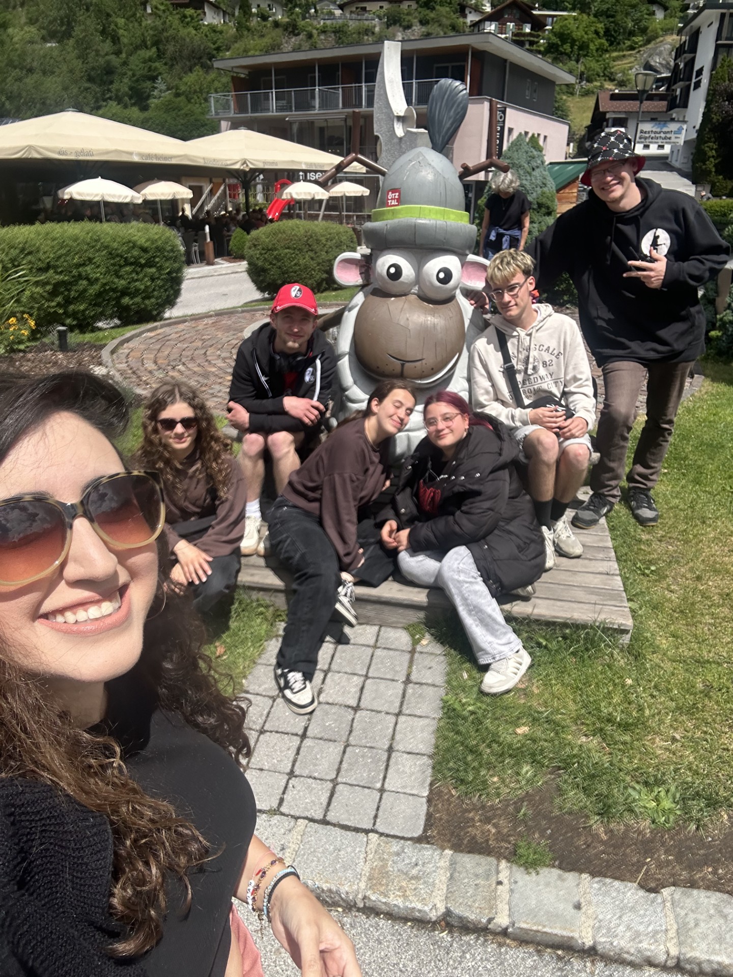 Selfie-Gruppe im Freien mit einer großen Comic-Figur in der Mitte.   Im Vordergrund links ist ein lachendes Gesicht einer Frau mit langen lockigen braunen Haaren, großer Sonnenbrille und schwarzem Oberteil, der Unterarm mit Armbändern ist zu sehen, sie hält die Kamera schräg von oben.   Hinter ihr sitzt und kniet eine Gruppe von sechs jungen Menschen auf einem gepflasterten Weg und auf einer niedrigen Holzbank, nah beieinander um eine große graue Tier-Figur herum.  