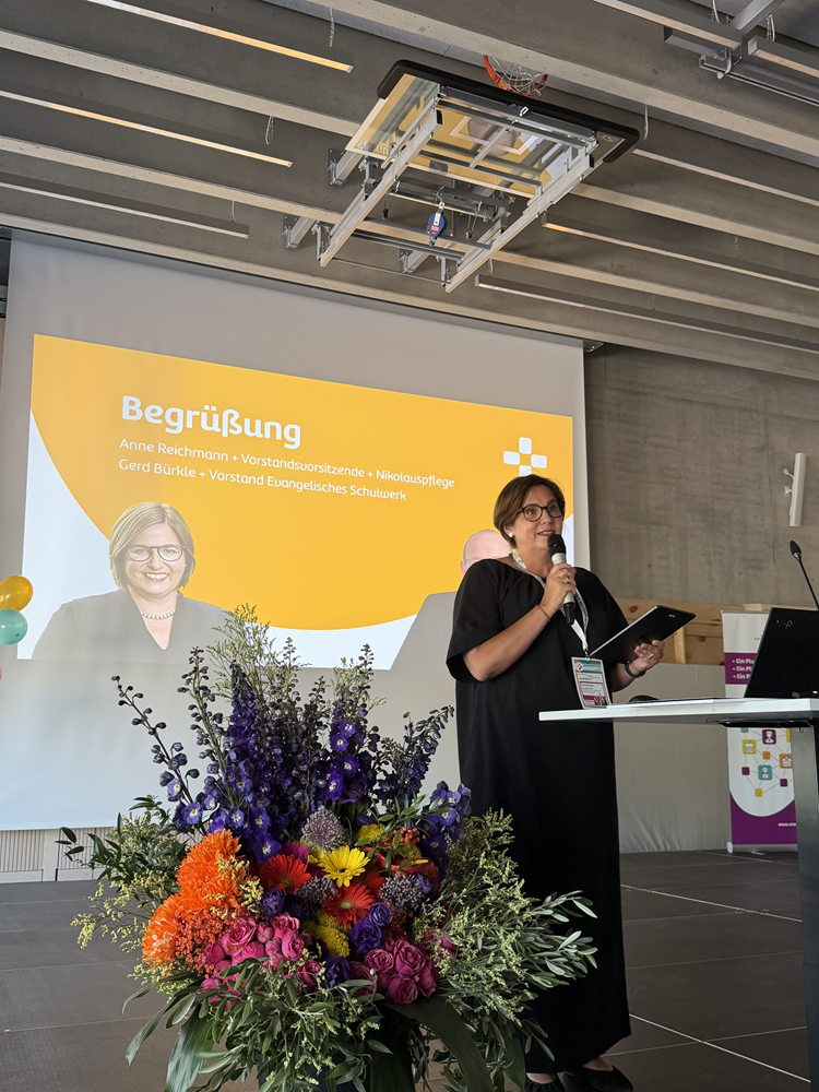 Auf dem Foto ist eine Frau auf einer Bühne an einem Rednerpult mit Mikrofon zu sehen, daneben ein großer bunter Blumenstrauß im Vordergrund. Hinter der Person hängt eine Leinwand mit einer gelb-weißen Präsentationsfolie, auf der links ein Porträtfoto derselben Person und darüber Text steht. Die Schrift in weiß lautet: „Begrüßung Anne Reichmann + Vorstandsvorsitzende + Nikolauspflege   Gerd Bürkle + Vorstand Evangelisches Schulwerk“. 